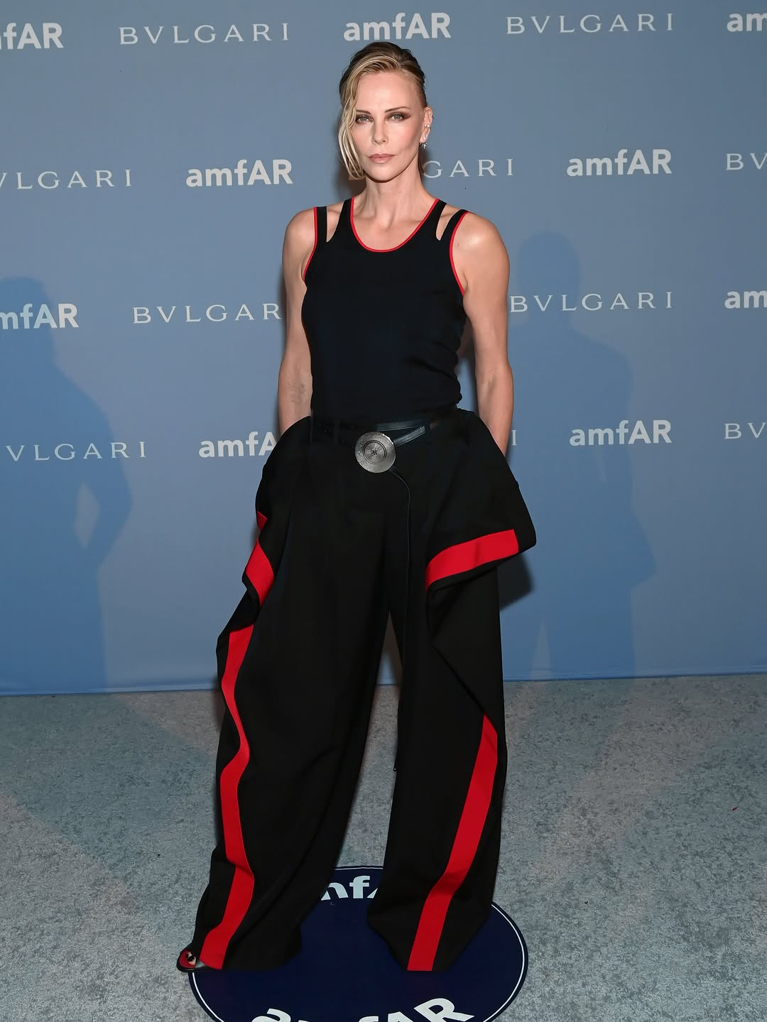 Charlize Theron, honoured with the Award of Inspiration, wore a black knitted sliced vest and wool pattern-piece trousers with the powder puff jewellery belt and Mobile zipped mules to amfAR Dallas 2026. Givenchy by S…